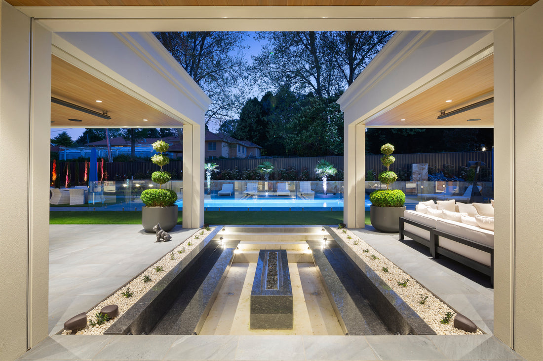 Covered outdoor entertaining area at dusk looking through to a lit pool, water feature and garden lighting